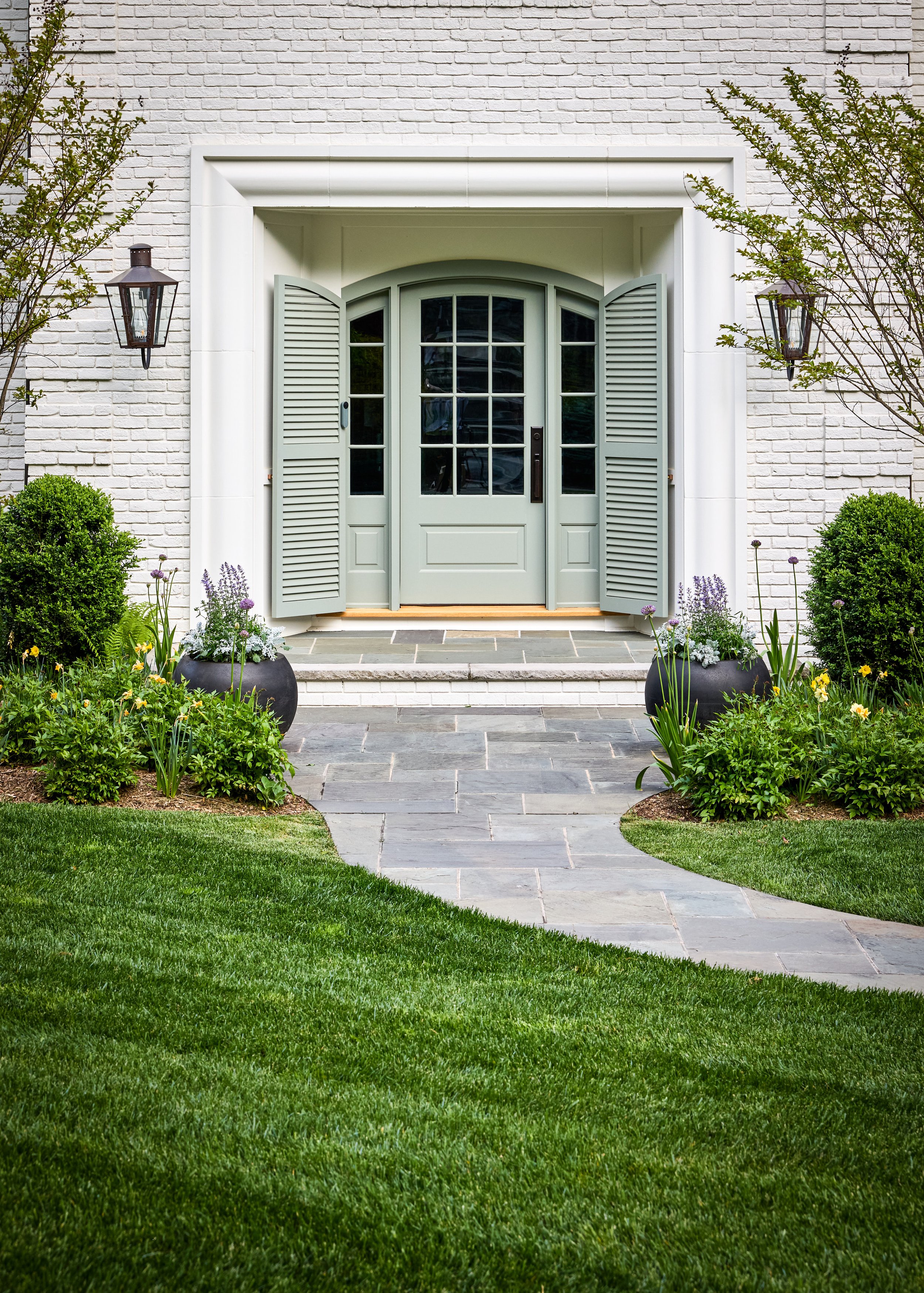 Arched sage green front door with louvered shutters, black gas lanterns, dark planters and a bluestone landing at a Mountainside, New Jersey home by Ellie Mroz Design.