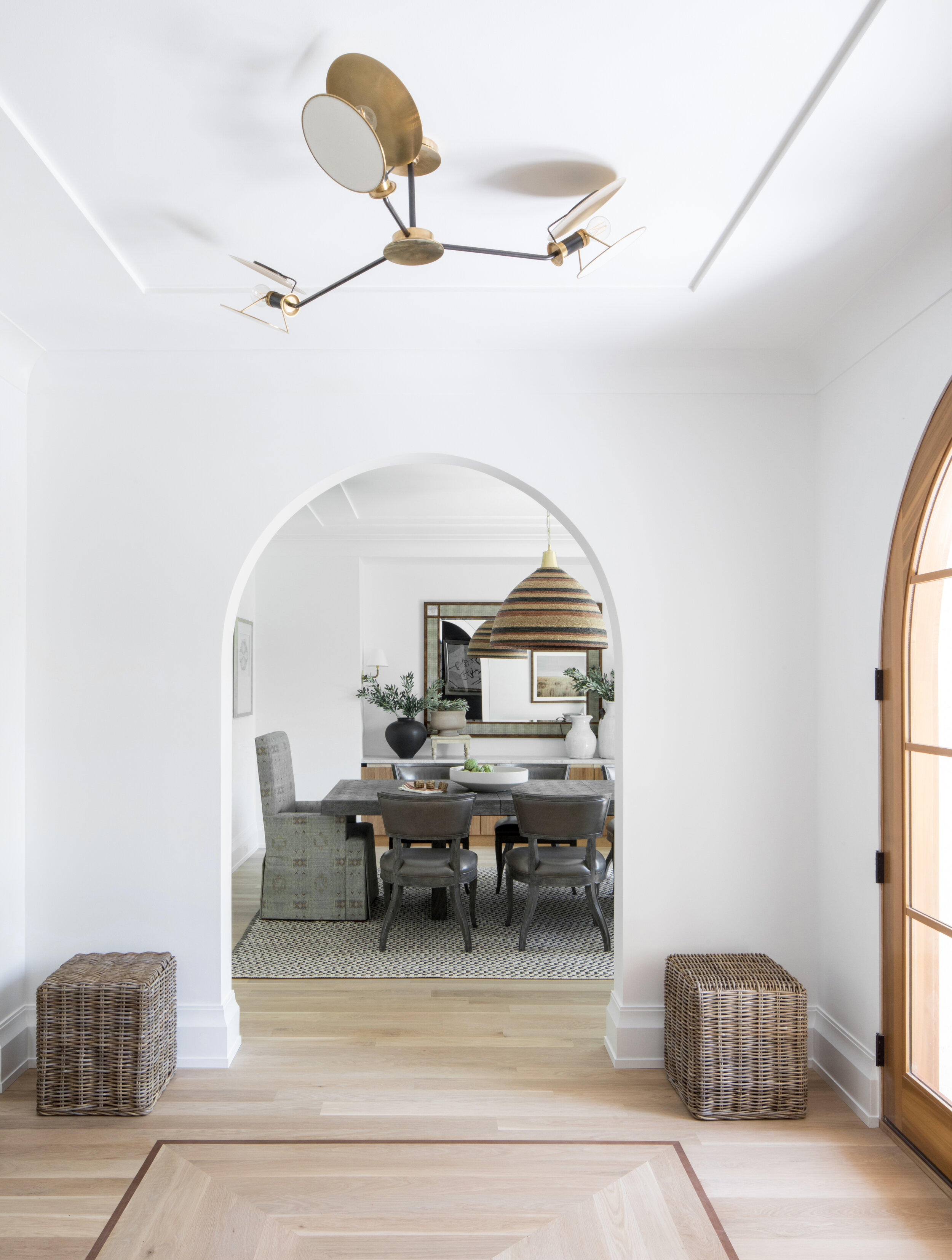 Entry with a sculptural brass ceiling light below a plaster arch, looking through to a dining room with a woven dome pendant, in a Westfield, New Jersey home by Ellie Mroz Design.