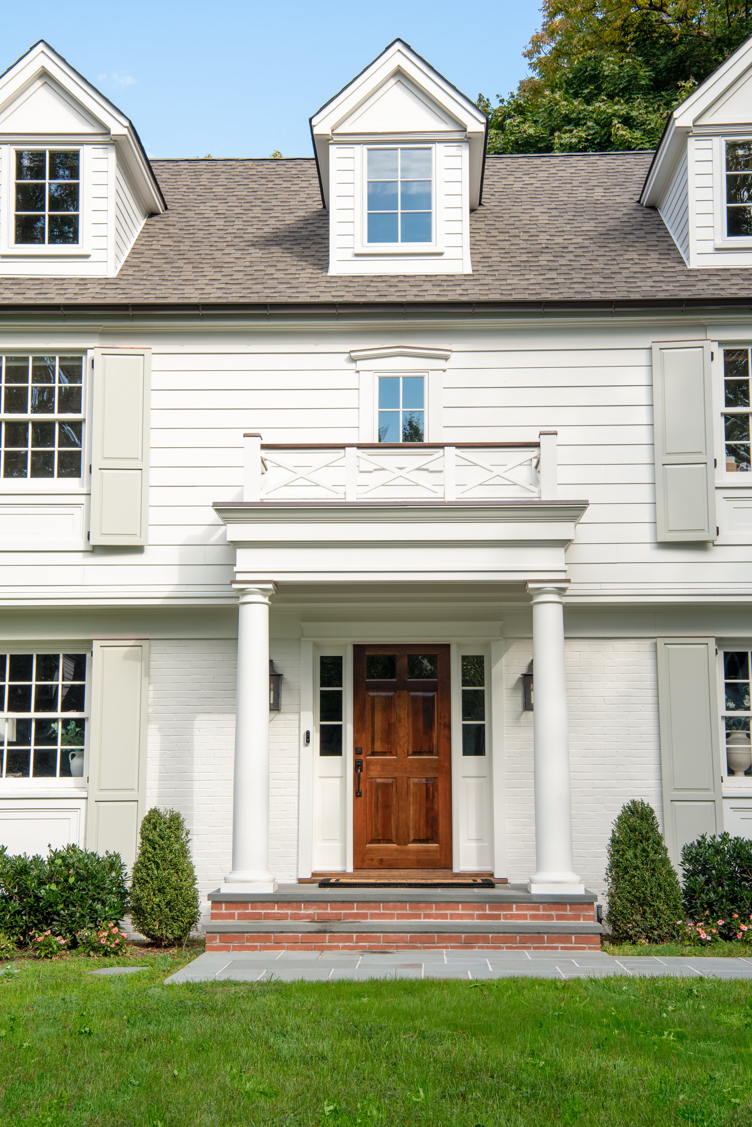 Columned portico with a stained wood front door and sidelights on a white clapboard colonial with dormers and sage shutters in Summit, New Jersey, by Ellie Mroz Design.