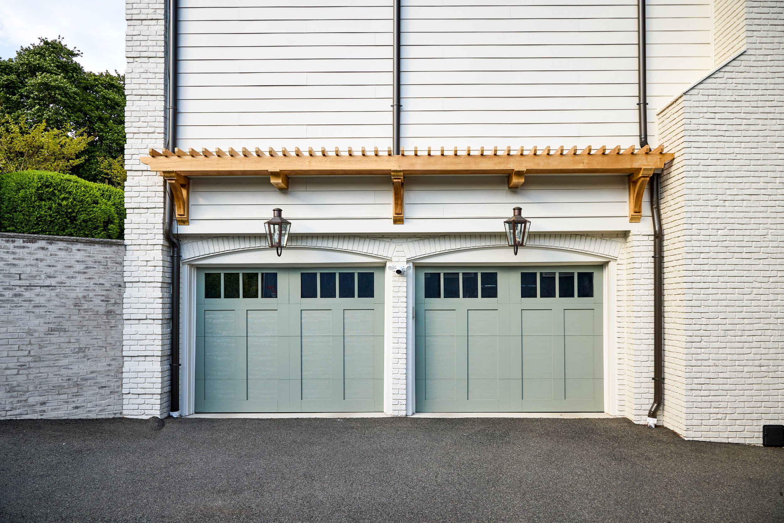 Sage green carriage-style garage doors under a cedar pergola with lanterns on either side, against white painted brick, at a Mountainside, New Jersey home by Ellie Mroz Design.