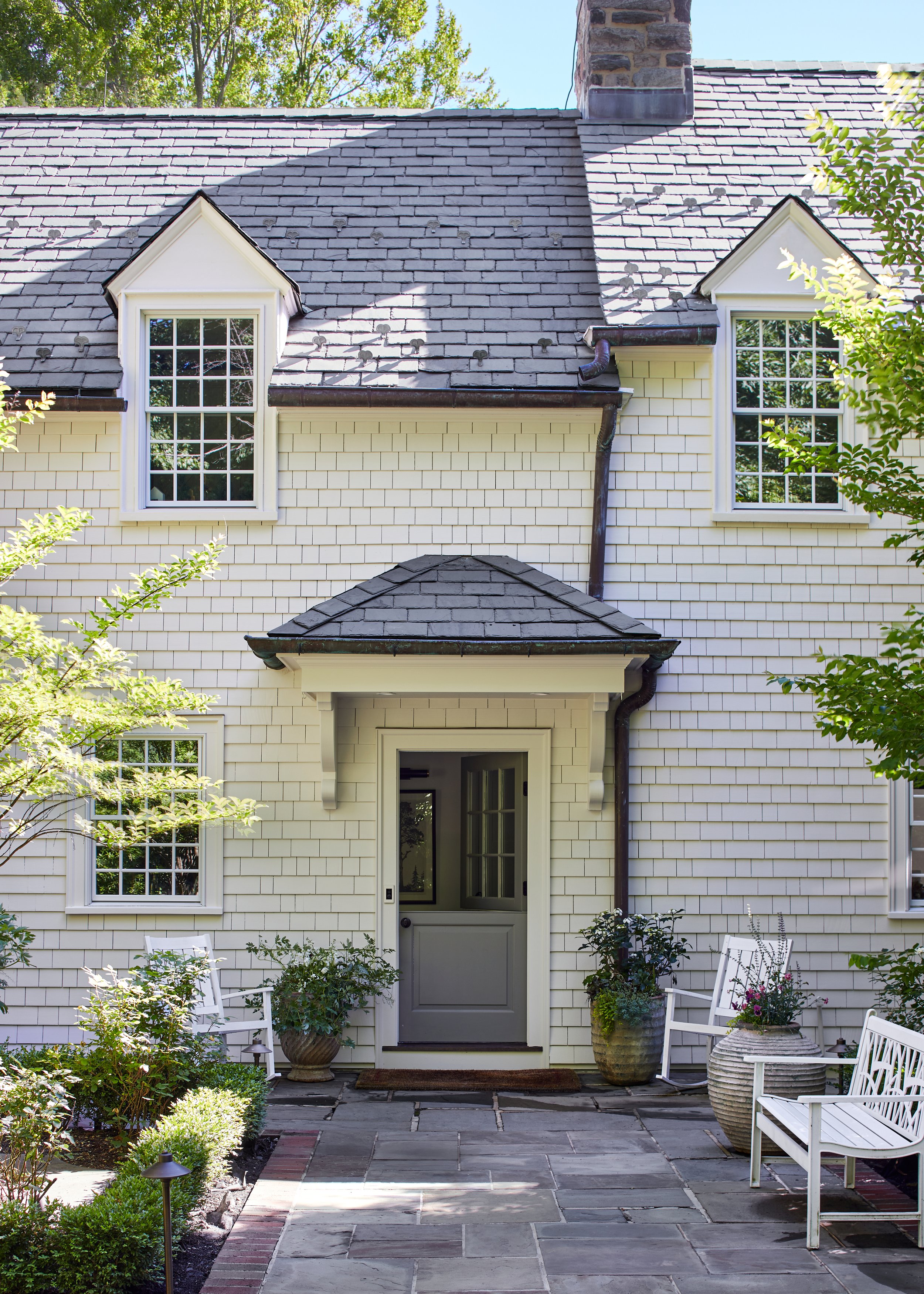 Cedar shingle home with a slate roof, white trimmed dormers and a dark entry door behind a planted front porch in Westfield, New Jersey, by Ellie Mroz Design.