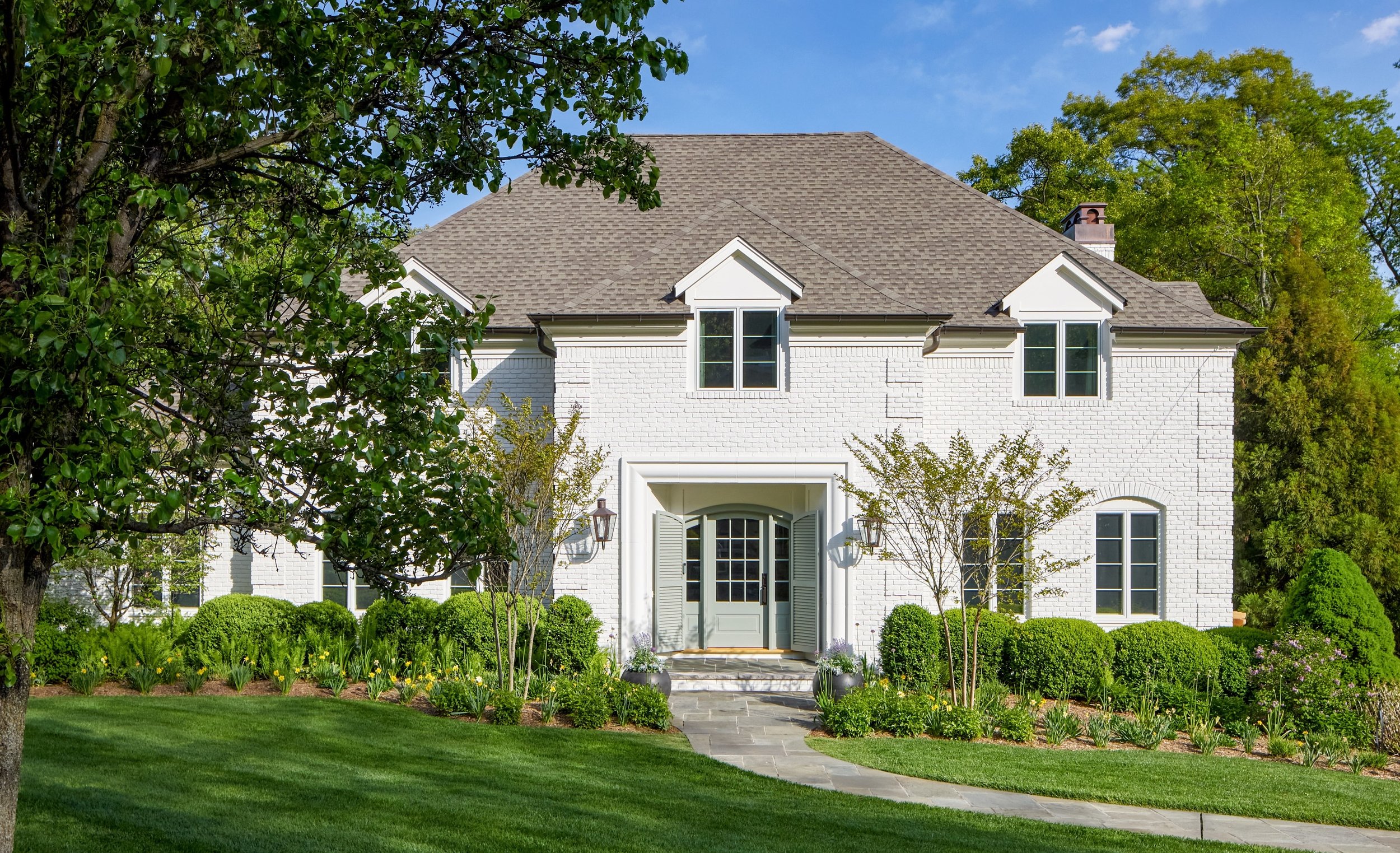 Front elevation of a white painted brick home with a steep slate-gray roof, dormers and clipped boxwood along a bluestone path, in a Mountainside, New Jersey renovation by Ellie Mroz Design.