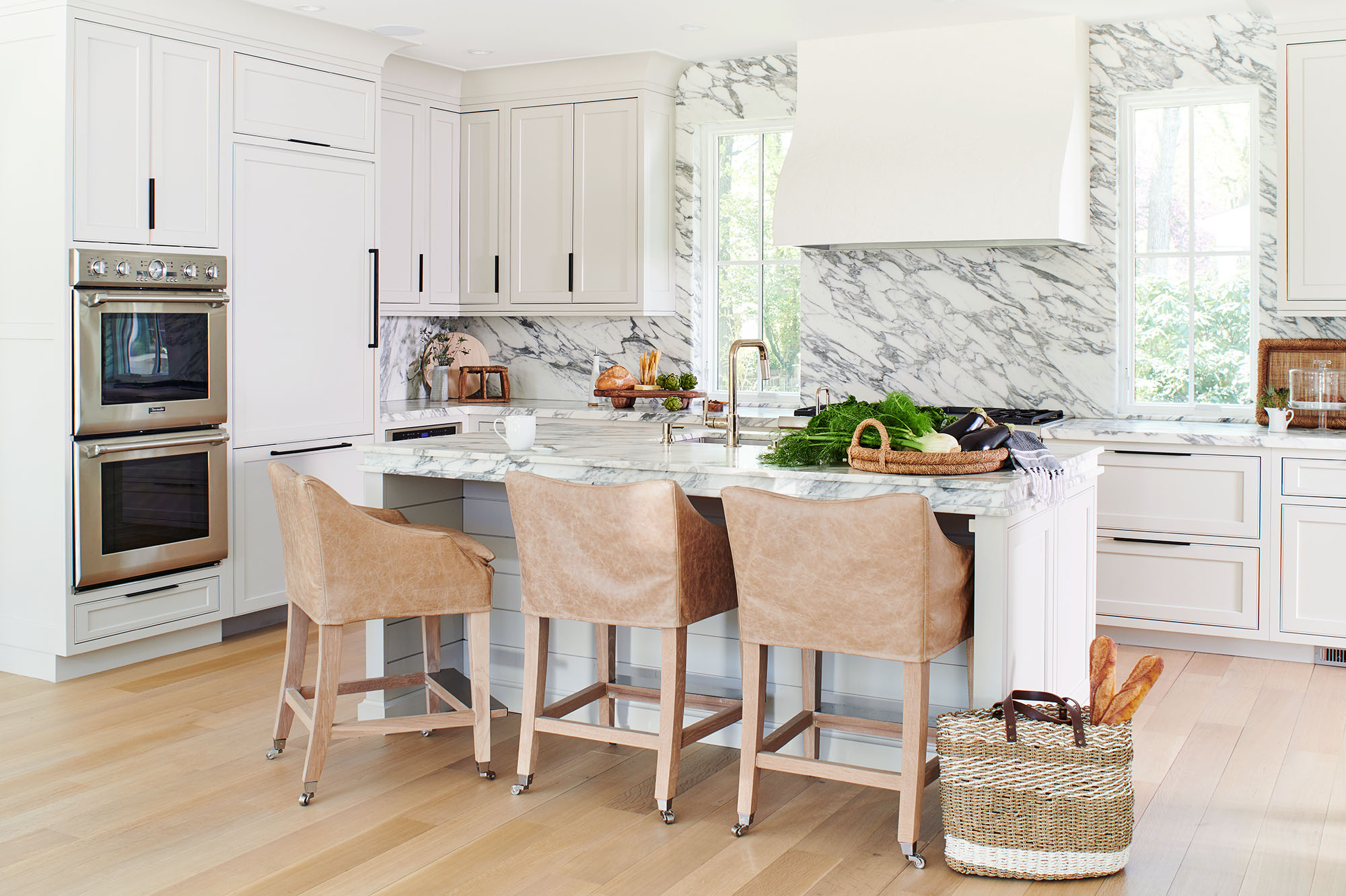 White kitchen with a marble island, tan leather counter stools and built-in wall ovens in a Westfield, NJ home by Ellie Mroz Design.