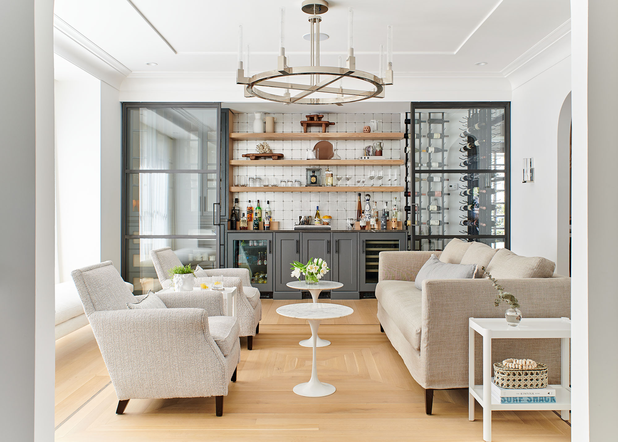 Bar room with a metal-and-glass wine wall, dark cabinetry, a round iron chandelier and cream armchairs in a Westfield, NJ home by Ellie Mroz Design.