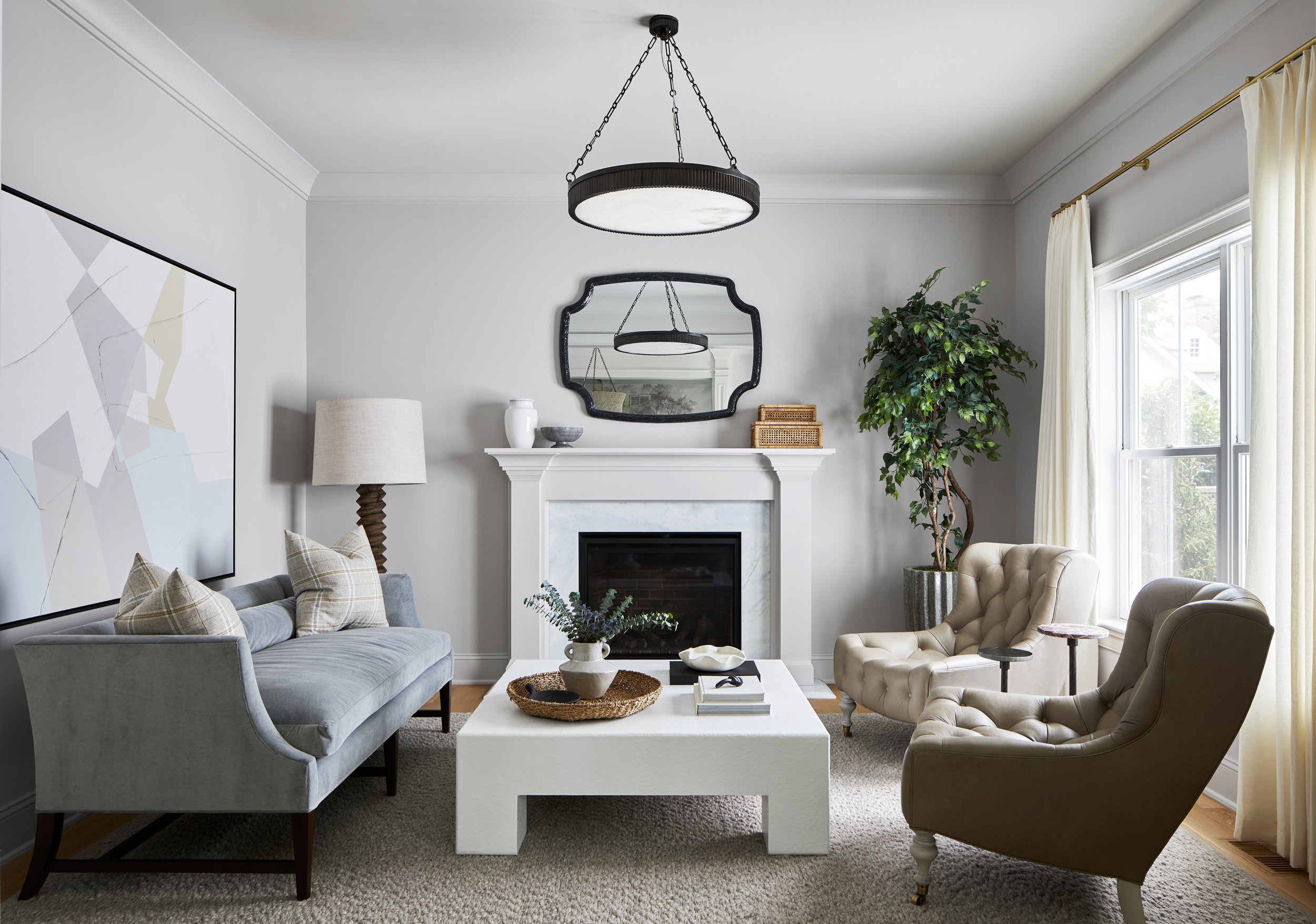 Living room with a traditional white fireplace, tufted wing chairs, a low white coffee table and a drum chandelier in a Chatham, New Jersey home by Ellie Mroz Design.