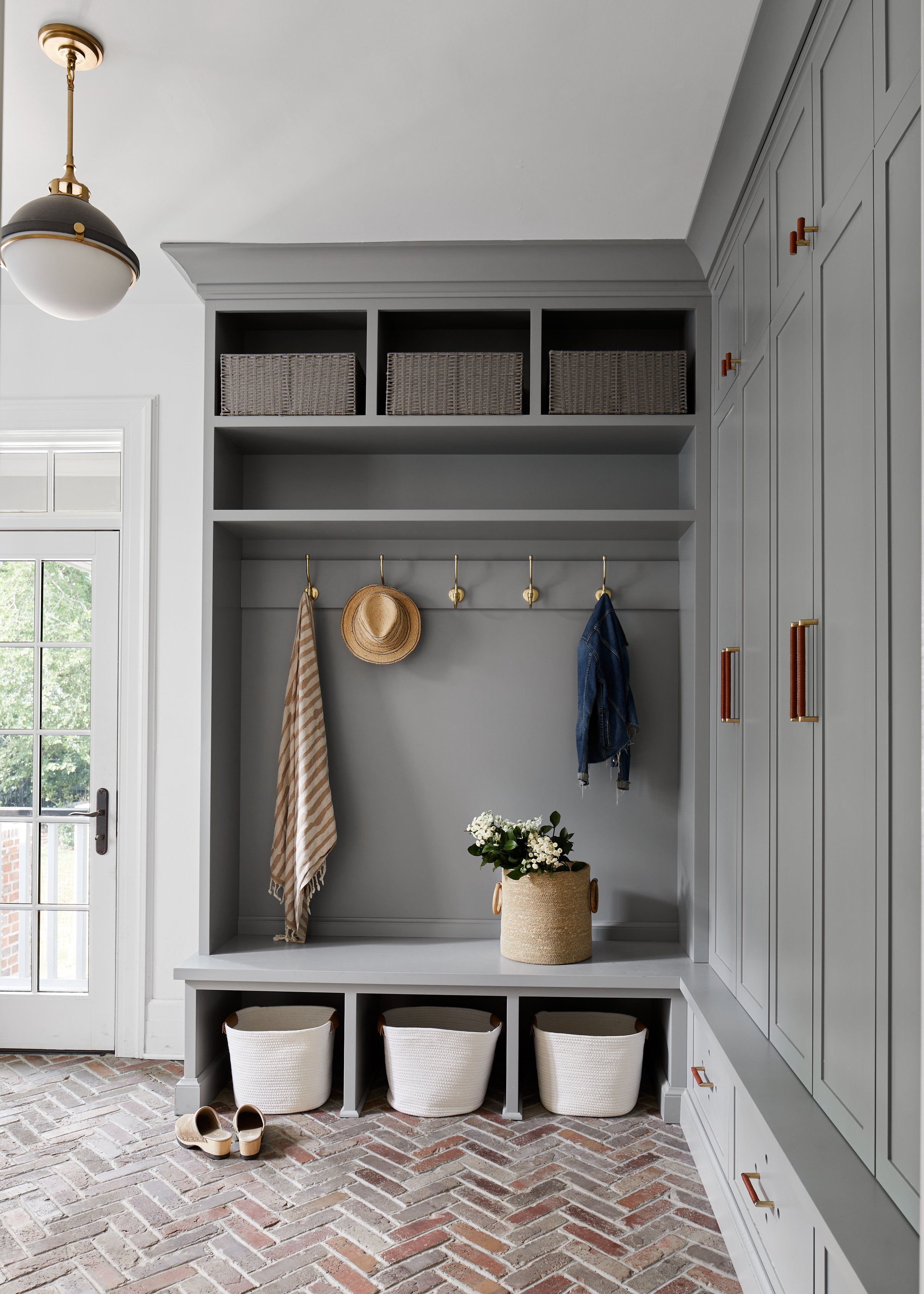 Blue-gray mudroom lockers with open upper cubbies, woven baskets, a row of brass hooks and white bins under the bench, above a herringbone brick floor, in a 1905 Westfield, New Jersey renovation by Ellie Mroz Design.