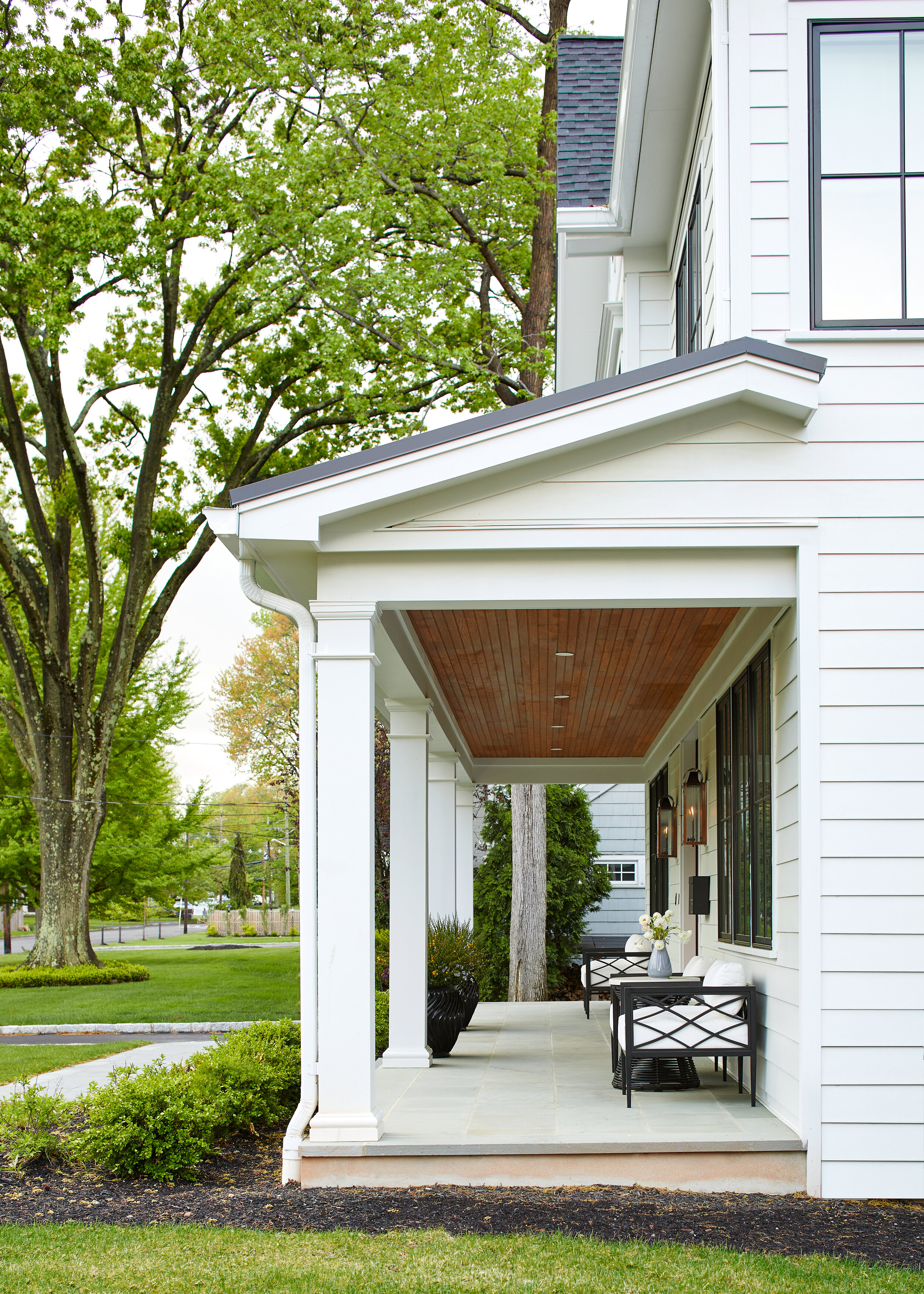A covered front porch with white columns, a stained wood ceiling and black wicker seating on a bluestone patio, on a Westfield, NJ home.
