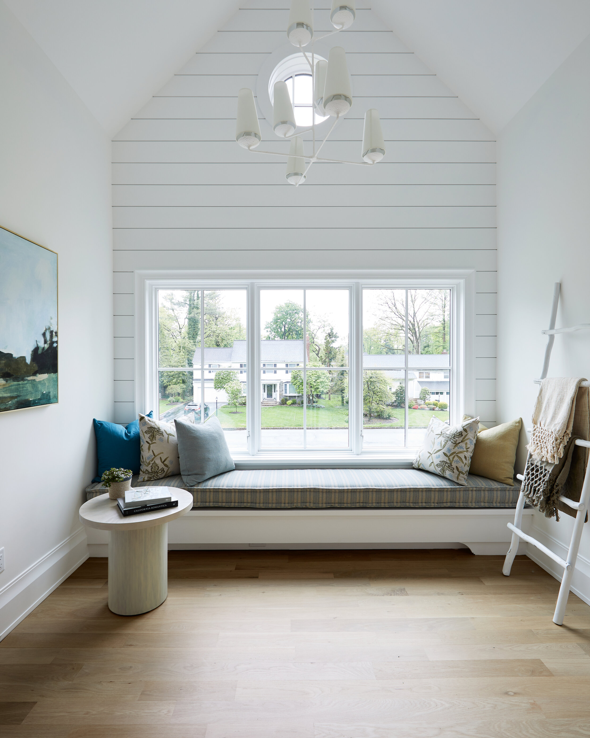A built-in window seat with a striped cushion and mixed pillows beneath a shiplap gable wall with a round accent window, in a Westfield, NJ home.
